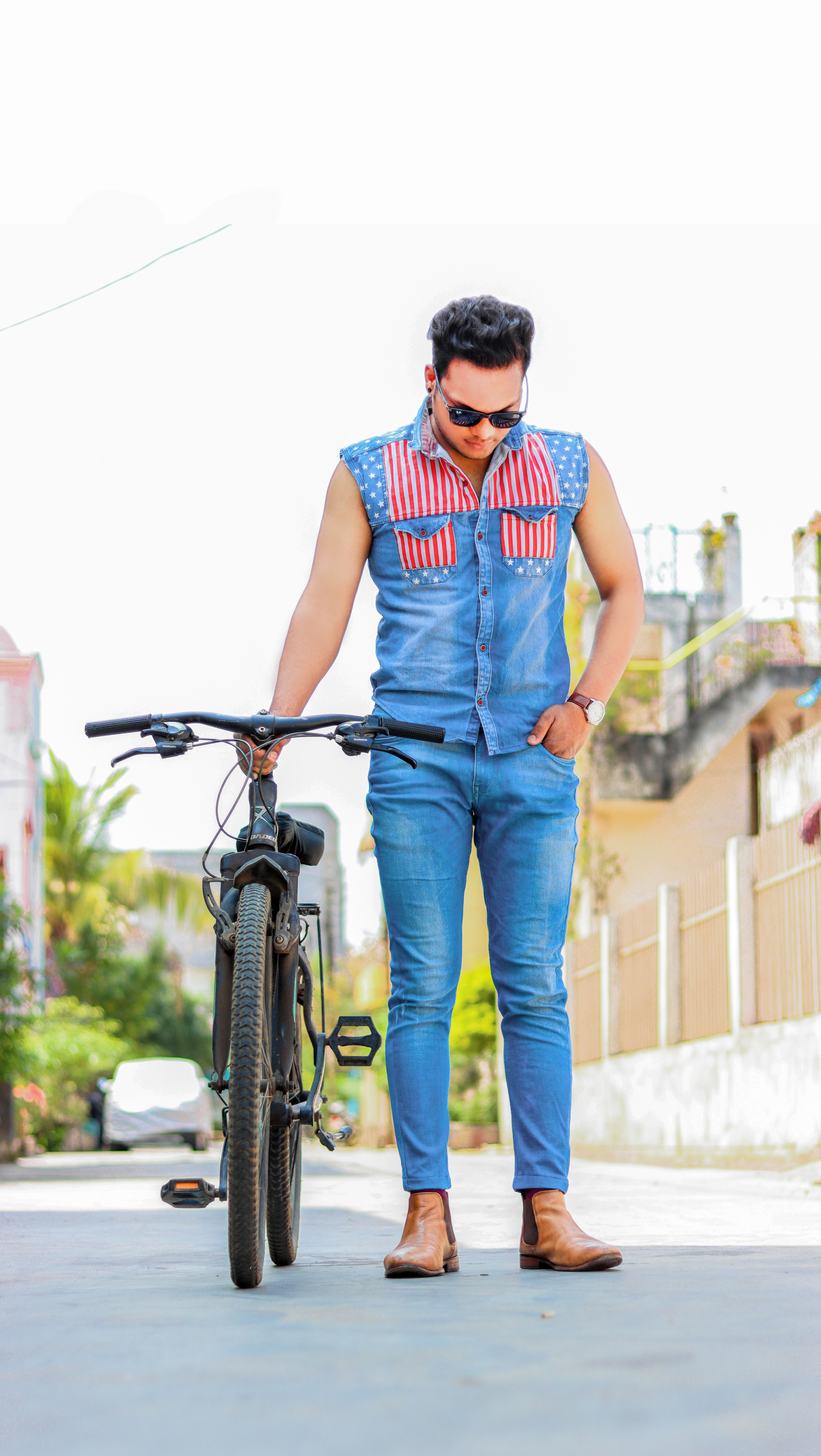 Stylish young man standing with a bicycle on a sunny city street, wearing a denim outfit and sunglasses.