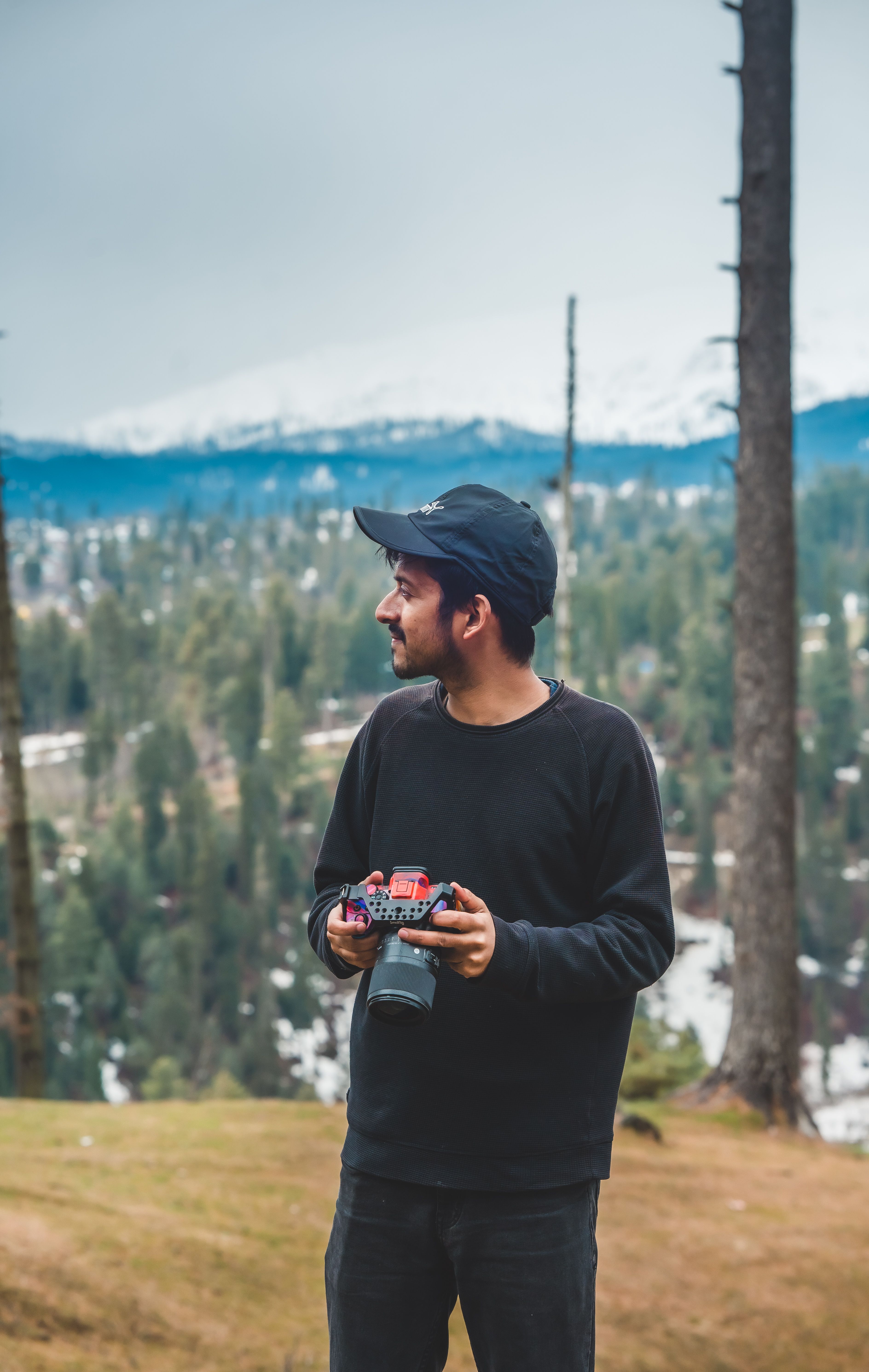 Man in a forest near Srinagar holding a camera. Snow-capped mountains in the background.