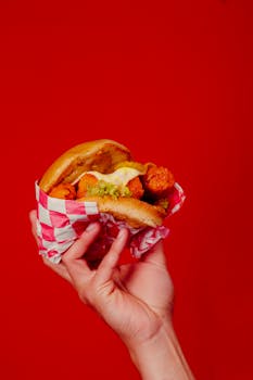 Close-up of a hand holding a delicious fried chicken sandwich wrapped in paper on a vibrant red background.