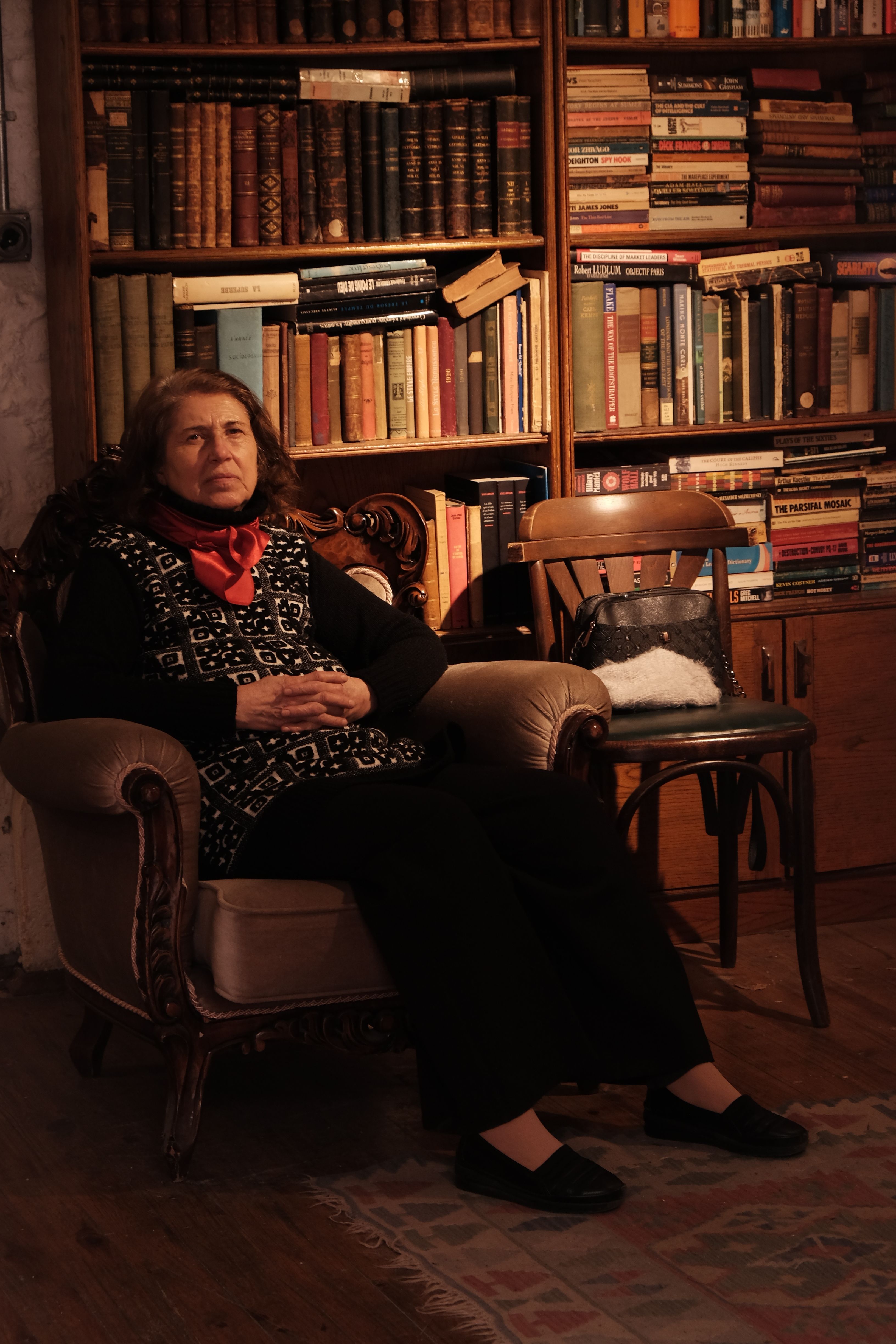 An elderly woman sits comfortably in an armchair within a cozy, vintage library room filled with books.