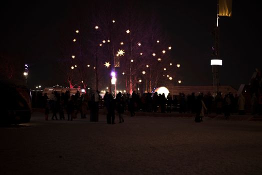 A cityscape at night featuring festive star-shaped lights and people gathering outdoors in winter.