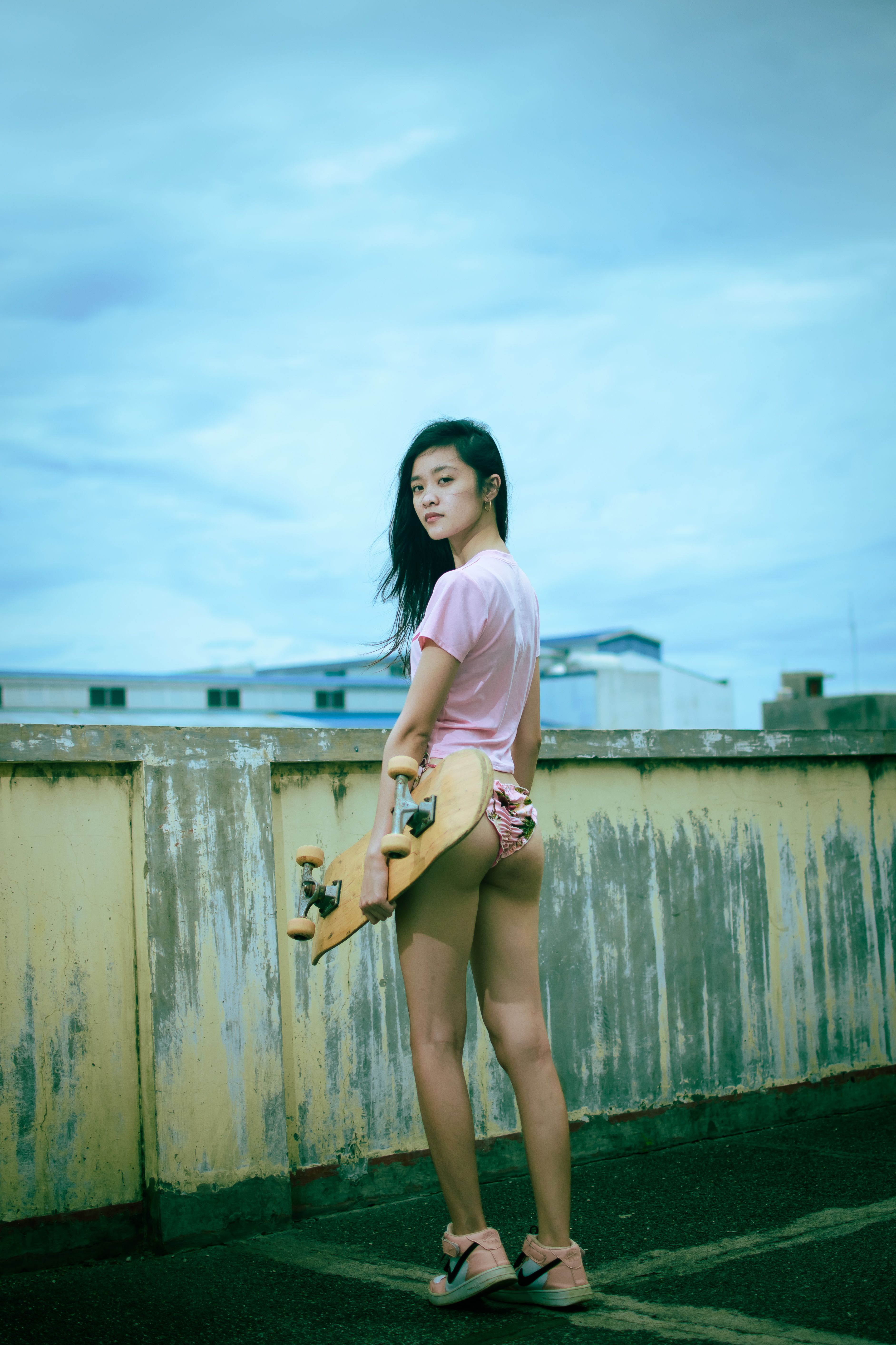 A young woman poses with a skateboard on a rooftop under a summer sky.