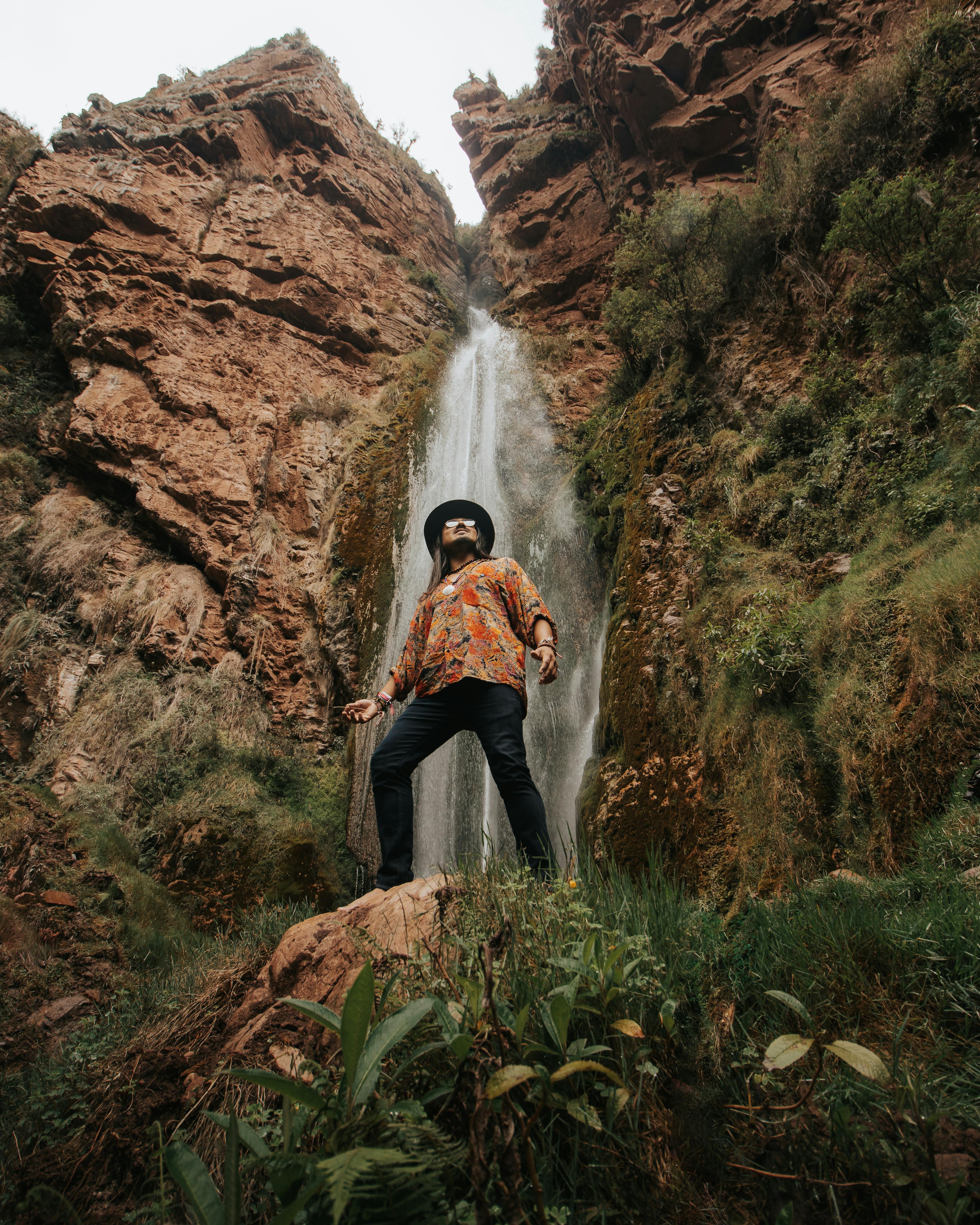 Captivating view of a young man in a vibrant shirt and hat, posing near a majestic waterfall.