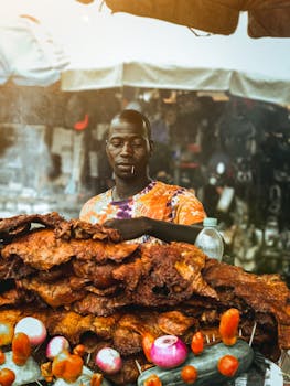 Man grilling meat at street stall with colorful vegetables in a bustling market.