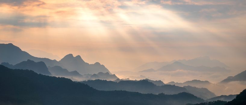 Peaceful mountain range at sunrise with sunlight streaming through clouds.