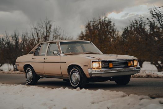 Classic car driving on a snow-covered rural road, under a dramatic winter sky.