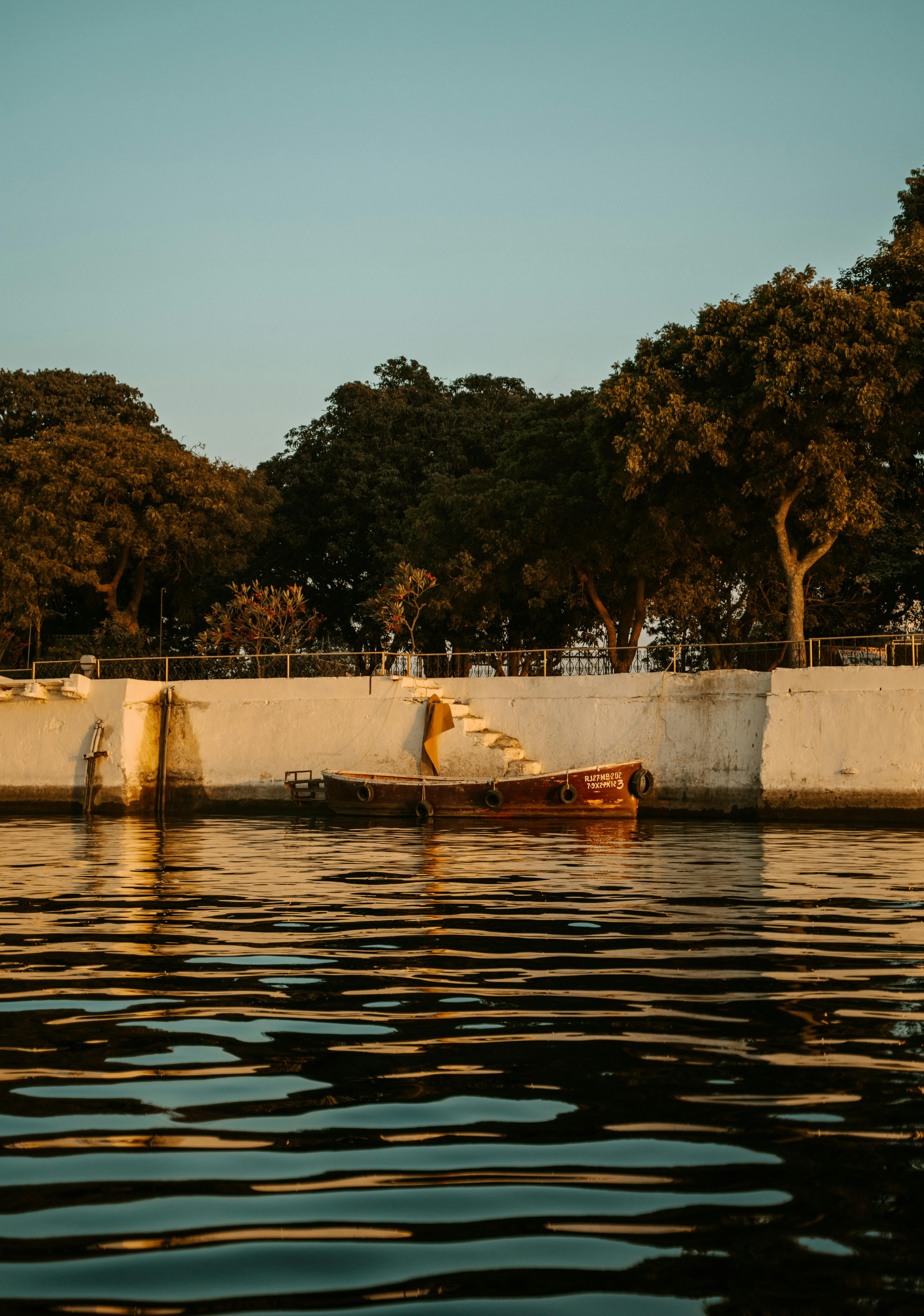 Peaceful scene of a wooden boat docked by a lakeside in India during golden hour with trees and clear sky.