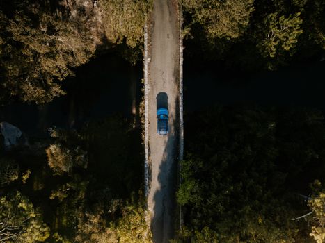 Top down drone shot of a blue car driving over a bridge surrounded by dense forest.