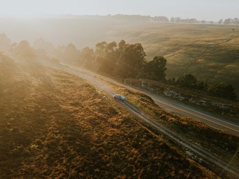 Aerial view of a car driving on a rural road surrounded by fields during sunrise.