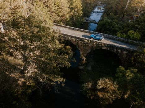 A blue car travels across a historic stone bridge spanning a forested stream, captured from above.