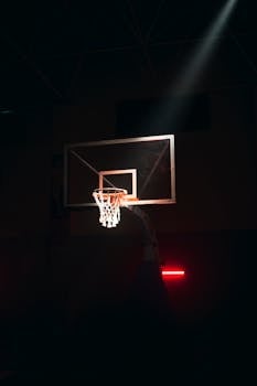 A basketball hoop highlighted by a spotlight in a dark indoor gym setting, creating a dramatic atmosphere.