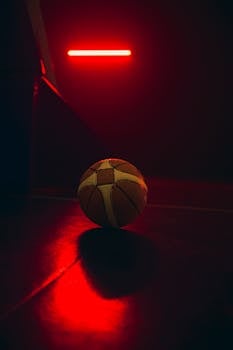 A basketball on a dark indoor court illuminated by a striking red neon light.