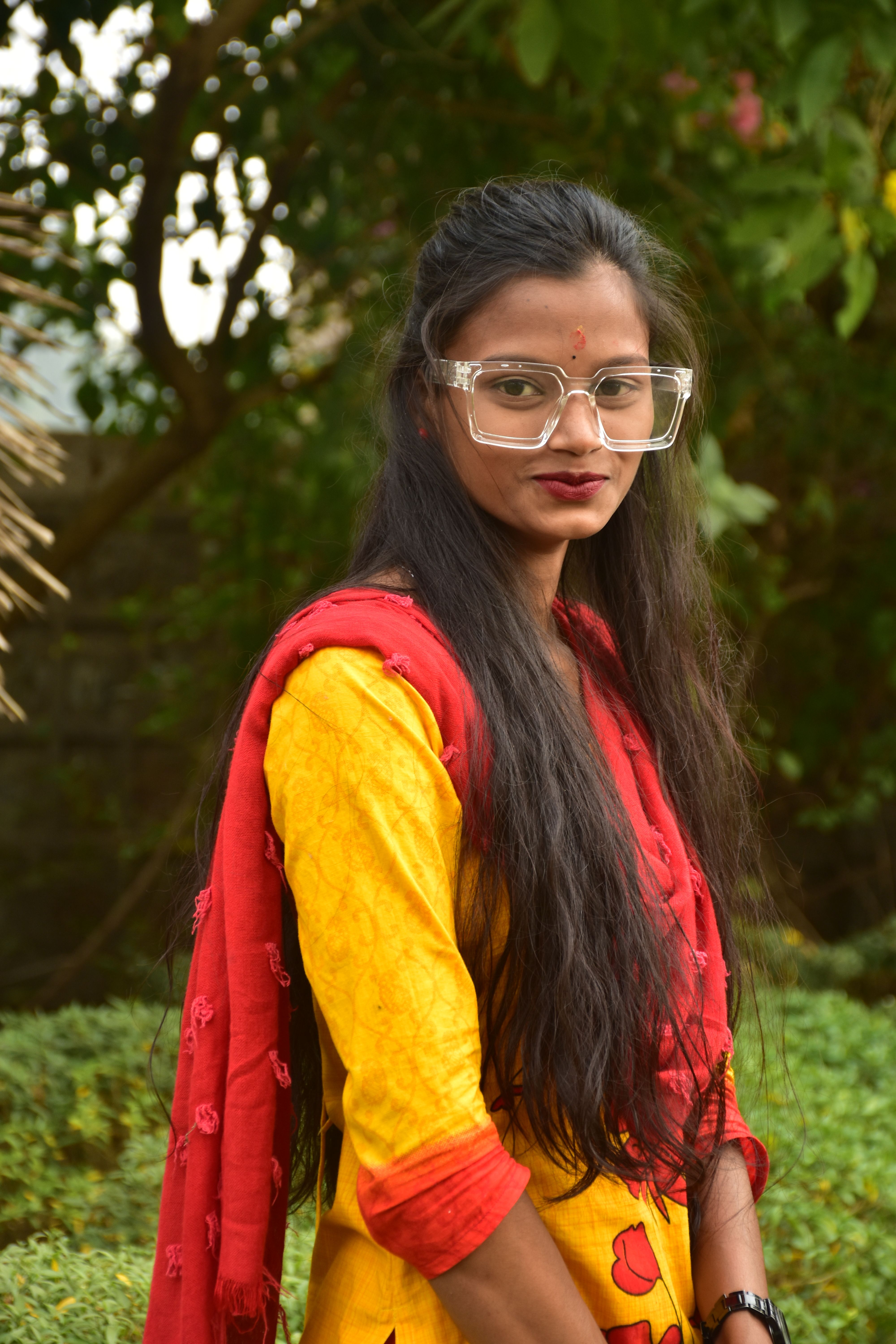 Portrait of a young woman wearing glasses and a colorful saree, smiling and standing outdoors among lush greenery.