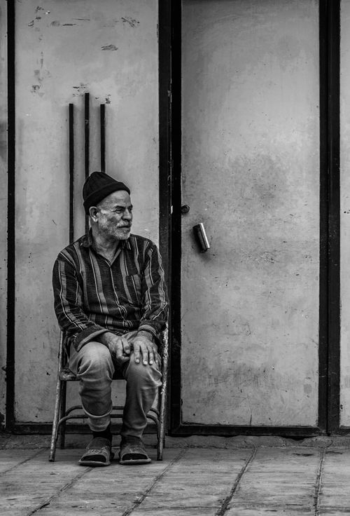 Free A black and white portrait of an elderly man sitting on a street against a wall. Urban scene. Stock Photo