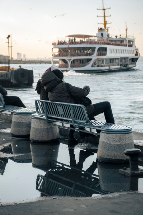 Free A couple enjoys a tranquil moment by the sea as a ferry passes by, reflecting the city's serene vibe. Stock Photo