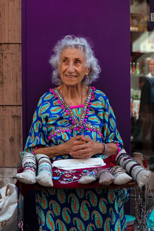 Free Smiling elderly woman selling handmade jewelry in a colorful urban setting. Stock Photo