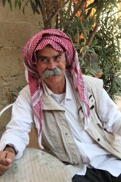 Eldery Man in an Arabic Scarf on his Head Sitting at an Outdoor Table 