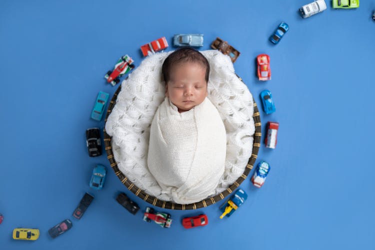 A Baby Boy Wrapped In A Nappy Lying On A Blanket On Blue Background With Toy Cars