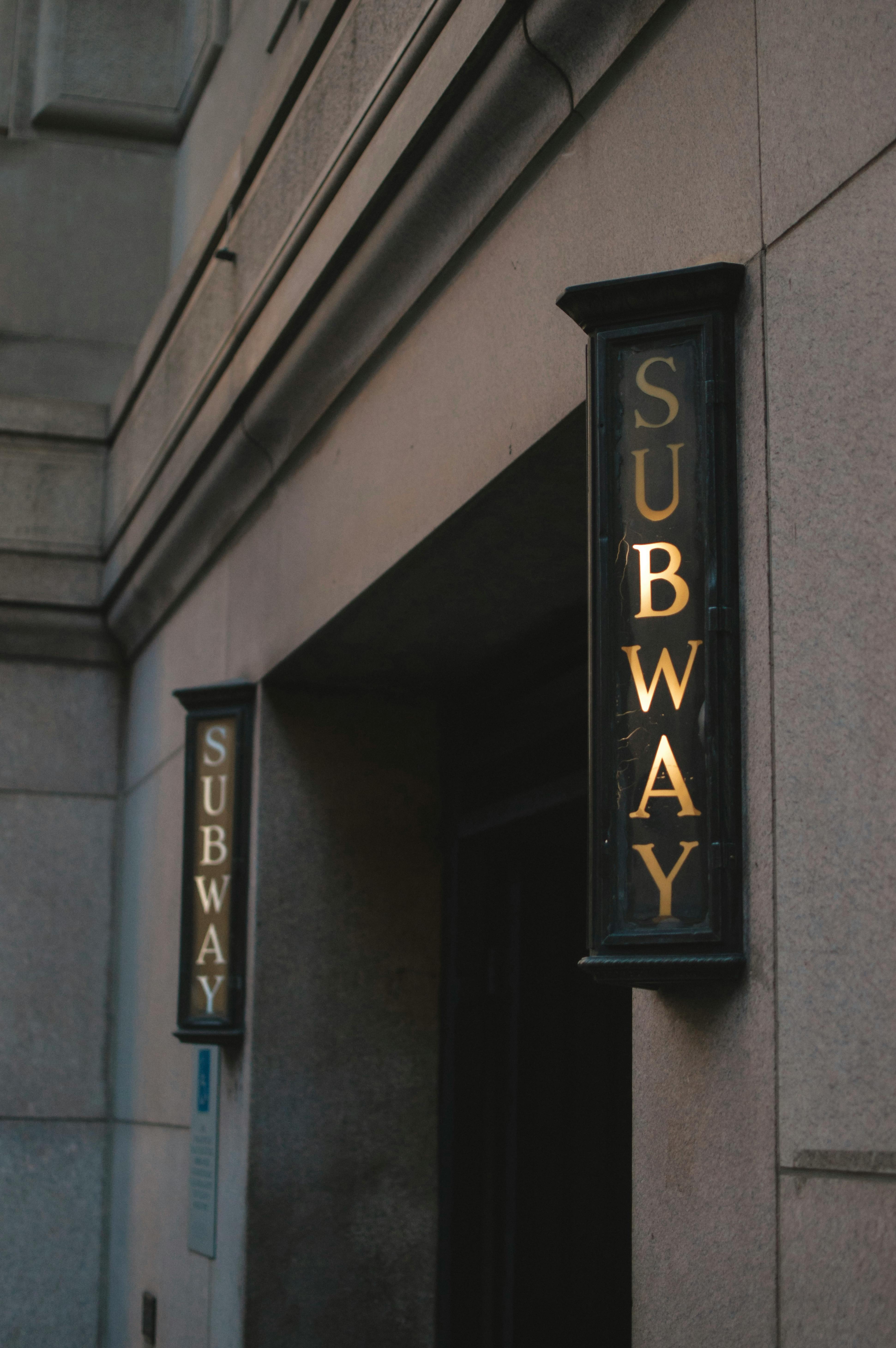 A classic subway entrance sign on a stone building exterior in an urban setting.