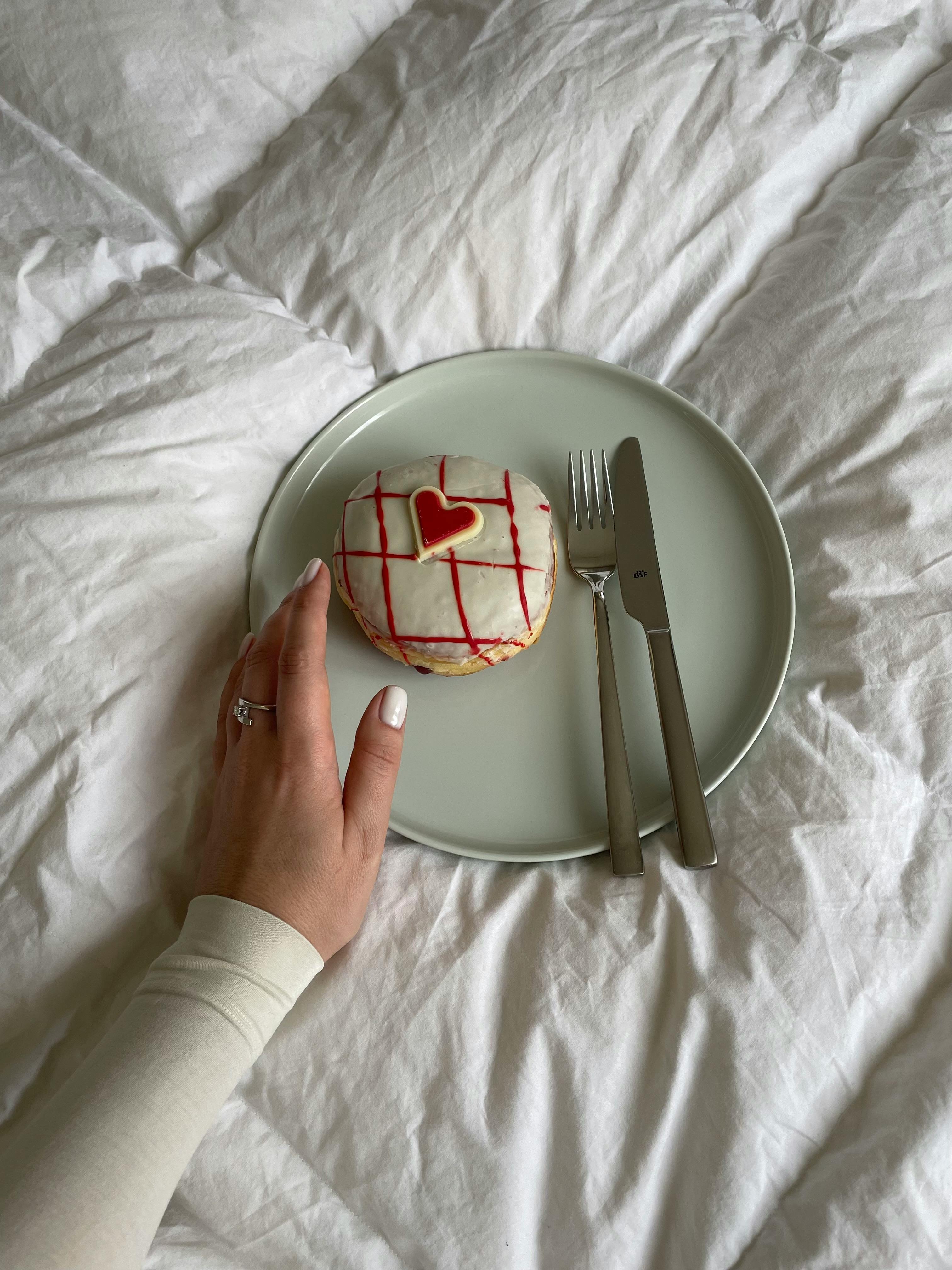 A hand holds a heart-decorated donut on a white plate with cutlery, offering a cozy and aesthetic visual.