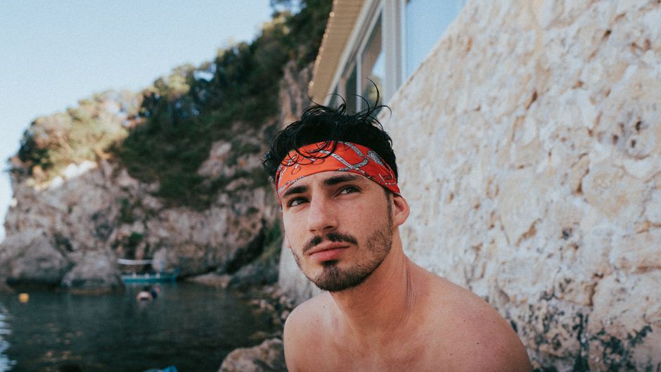 A shirtless young man with a red bandana poses by the beautiful rocky shores of Kerkyra, Greece.
