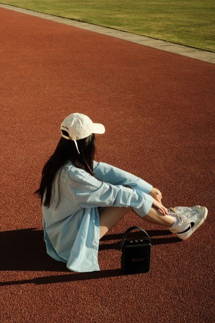 A woman in casual fashion sits on a track wearing a cap and sneakers, enjoying leisure time.