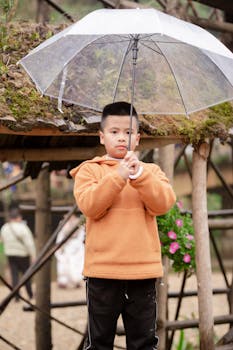 Child standing outdoors in casual orange hoodie holding a transparent umbrella on a cloudy day.