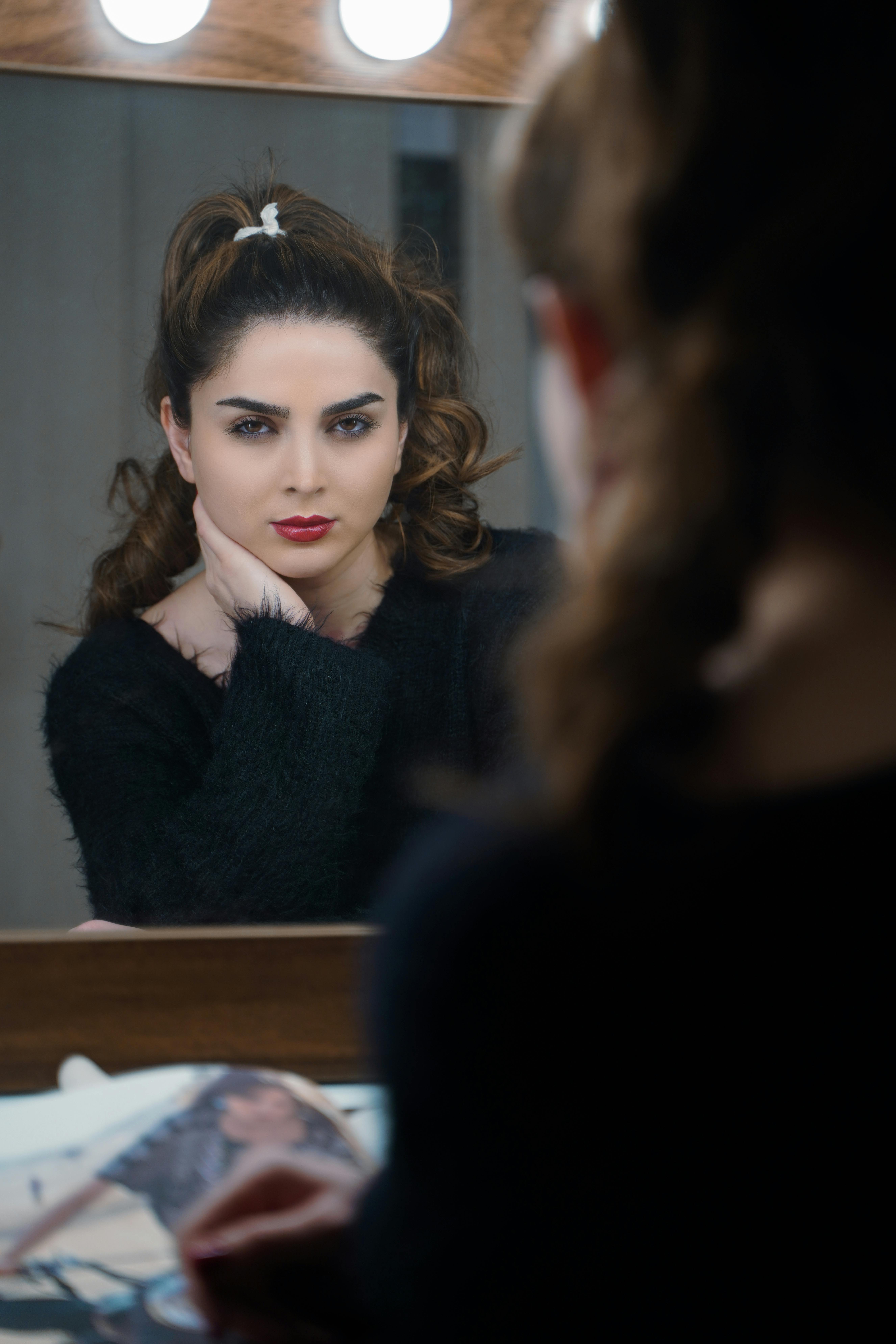 A woman with red lipstick looks into a mirror, reflecting on her beauty in a vanity scene.