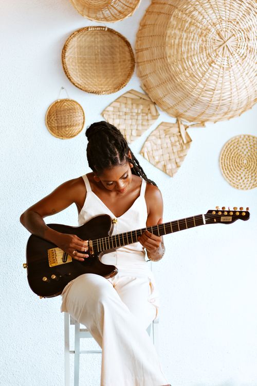 Free A young woman skillfully playing guitar against a white wall adorned with wicker baskets. Stock Photo