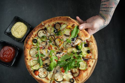 Free Top view of a vegetarian pizza with fresh toppings and sauces, held by a tattooed hand. Stock Photo