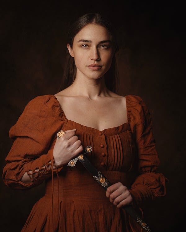 Model in a Medieval Square Neck Brown Dress With Laced Cuffs Drawing a Dagger From its Sheath