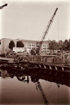Sepia-toned image of a construction site at a harbor with cranes and reflections on water.