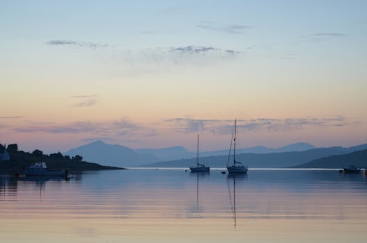 Tranquil sunset view with sailboats and mountains reflecting on calm water.