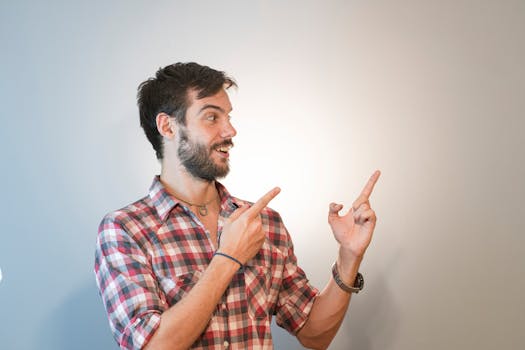 A young man in a checkered shirt smiling and pointing upwards, gesturing excitement in a studio setting.