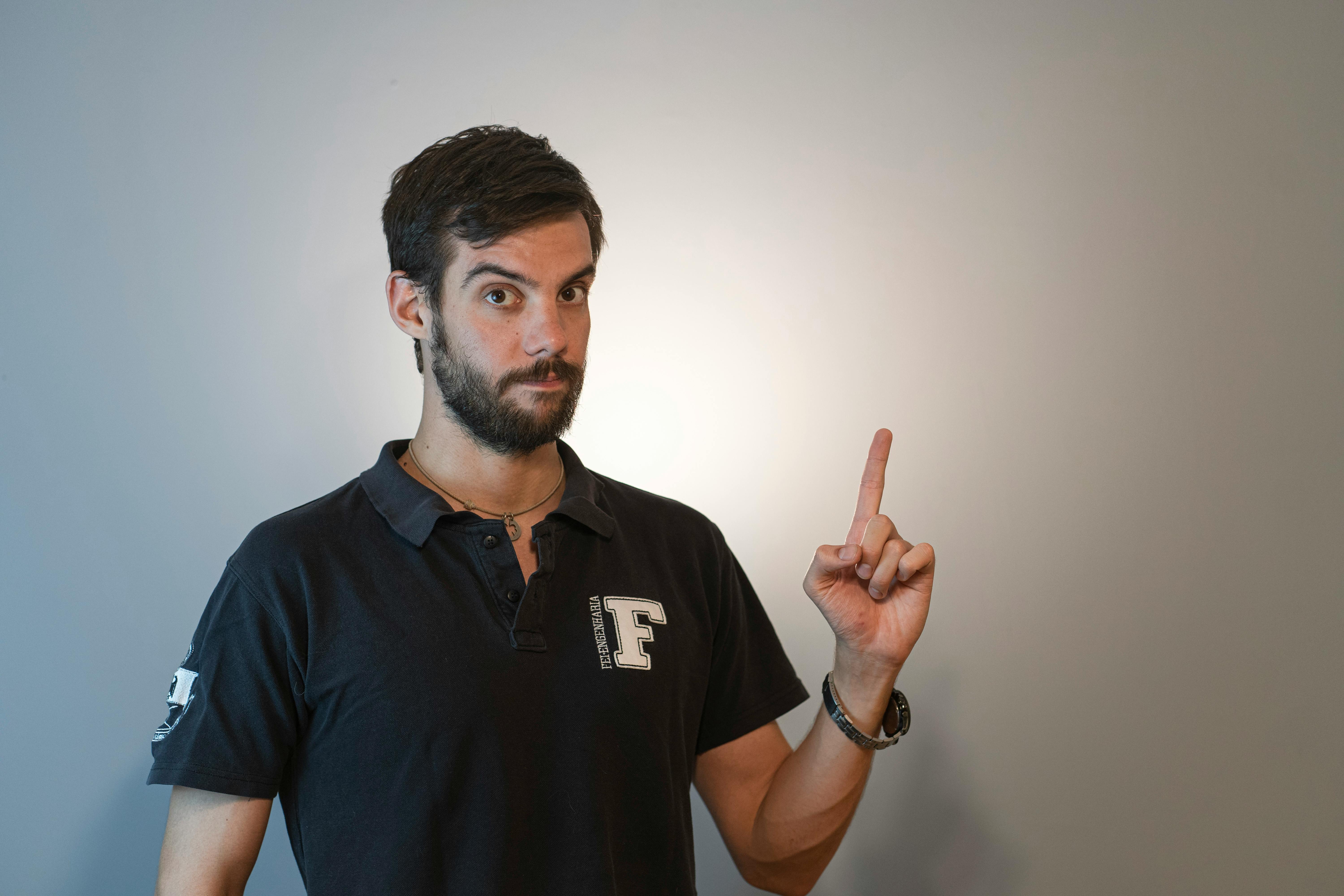 Bearded man in a polo shirt pointing upward against a plain studio backdrop.