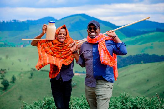 Two men in colorful shawls smiling and posing with a milk jug in open green fields.