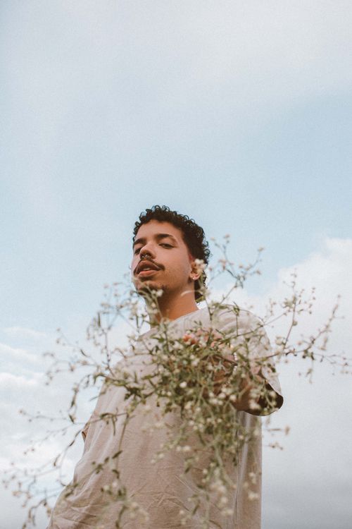 Free A young man holds wildflowers with a serene expression in an outdoor summer setting under a clear blue sky. Stock Photo