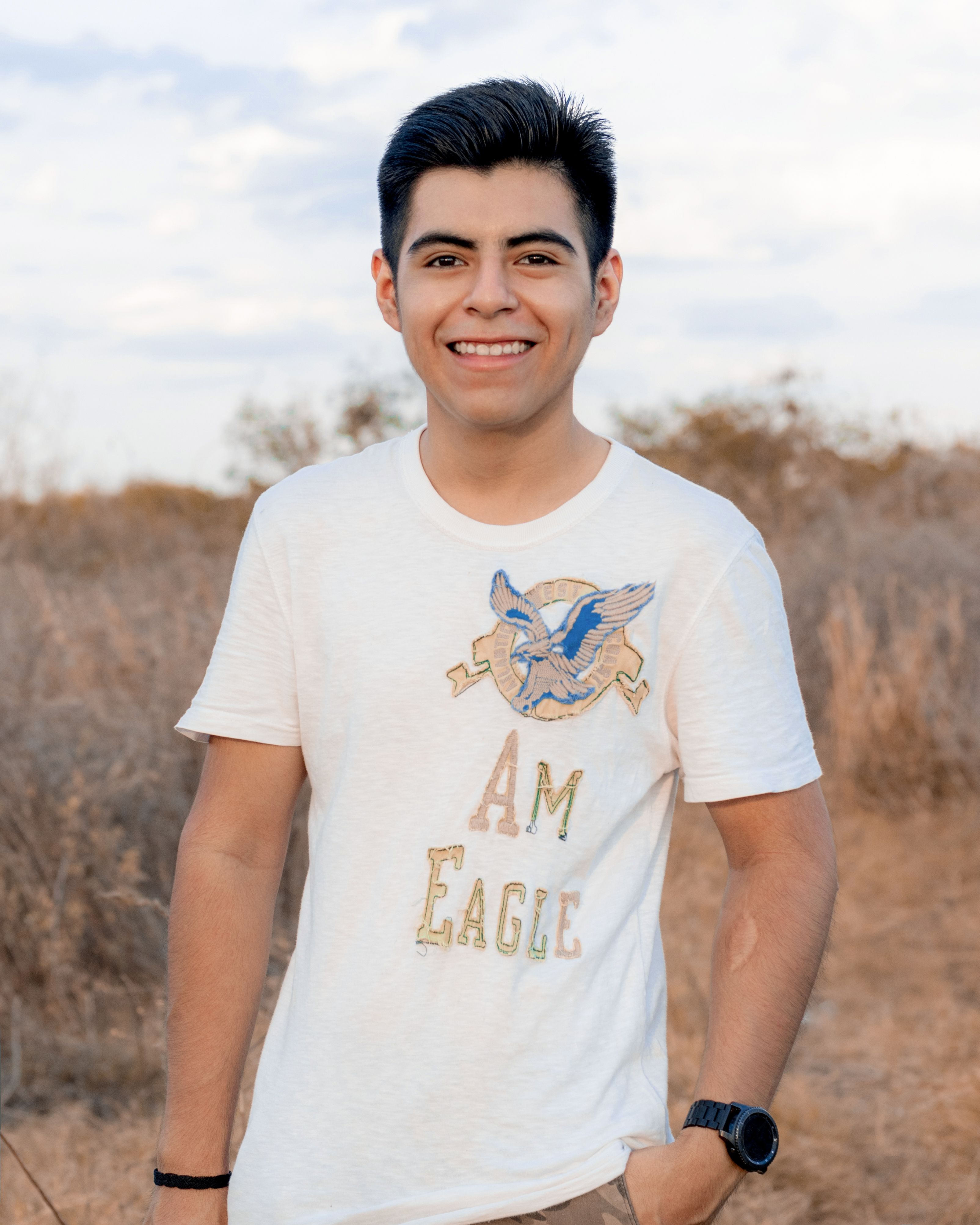 A young man smiling outdoors in casual attire with natural background.