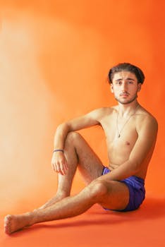 A topless young man with short hair poses on an orange background, creating a vibrant and striking scene.