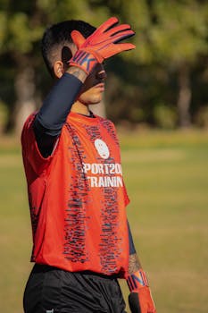 A goalkeeper in red sportswear shields eyes from the sun during outdoor soccer training.