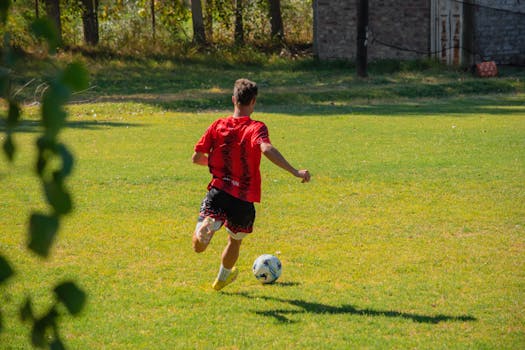 Back view of a male soccer player in action on a sunlit field wearing a red jersey.
