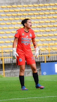 A female goalkeeper in bright orange uniform stands on the soccer field during a match.