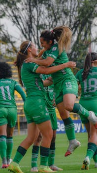 Joyful women's soccer team celebrating a victory on the field in green uniforms.