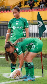 Two female soccer players in green uniforms preparing a corner kick on the field.