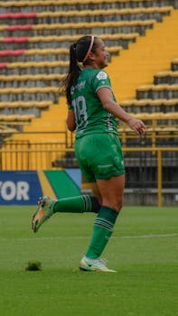 A female football player in a green uniform runs on a grass field in a stadium.