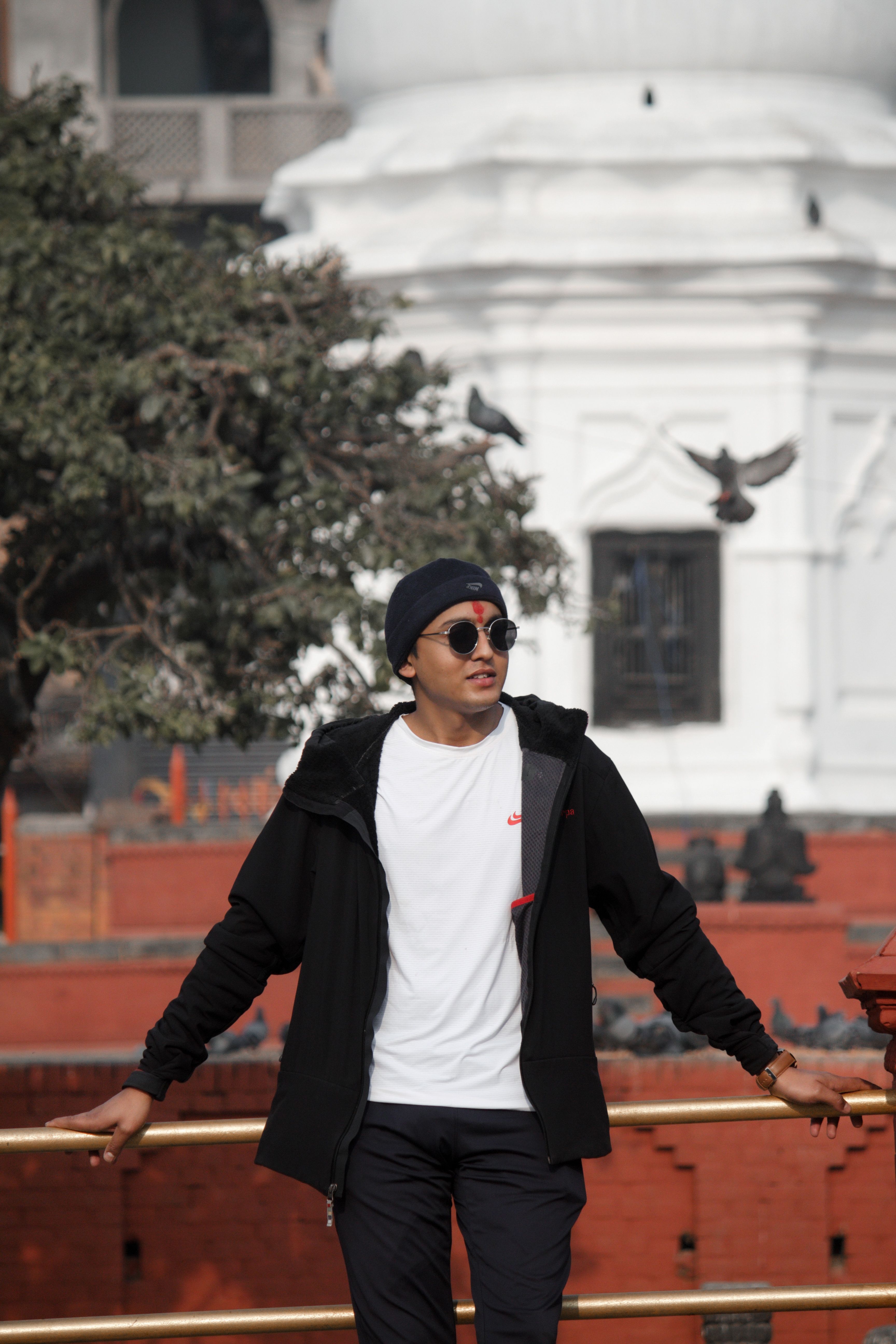 Young man in casual attire posing with sunglasses in an urban outdoor setting.