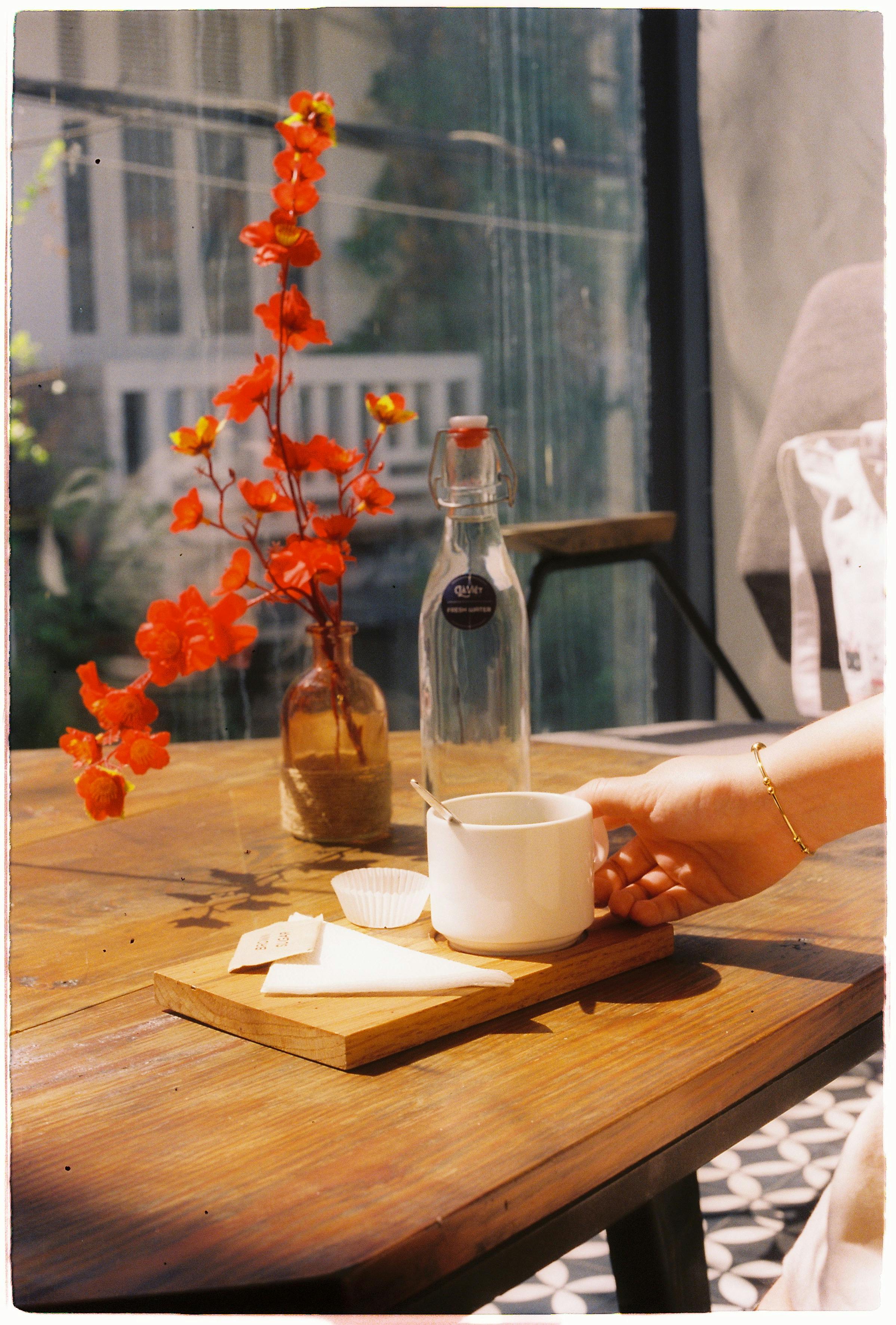 A serene cafe table setup with coffee, water bottle, and vibrant flowers in sunlight.