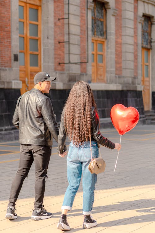 Free A Couple Walking on a Sidewalk while Holding Hands and Woman Holding a Heart Shaped Balloon  Stock Photo