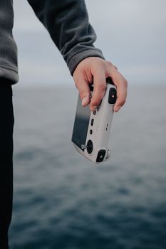Close-up of a person's hand holding a white drone controller by the ocean.