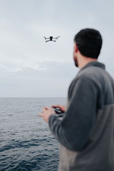 A man controls a flying drone over the ocean, showcasing modern technology and hobbyist interests.